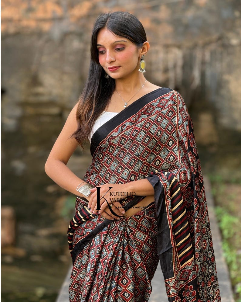 Pu&ograve; includere: Un sari nero caratterizzato da un motivo geometrico ripetitivo di quadrati rossi, bianchi e verde acqua con un bordo nero. Il tessuto presenta un design stampato con il logo KUTCH IQ VARS A BANJARA COUTURE visibile sulla parte drappeggiata.