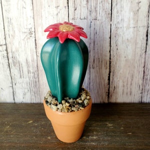 May include: A teal-colored cactus sculpture with a red flower and gold center sits in a small terracotta pot filled with pebbles. The cactus has a smooth, glossy finish. The background is a light-colored wooden surface.