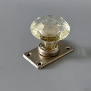 May include: A clear glass doorknob with a circular pattern, mounted on a rectangular metal backplate. The knob has a yellowish tint and a rounded shape. The metal backplate has a brushed finish and four screw holes.