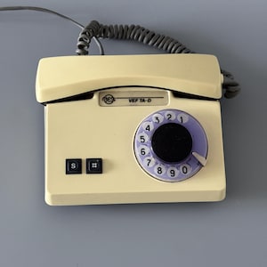 Puede incluir: Teléfono vintage color crema con un dial giratorio y un auricular. El dial es morado con números blancos. El teléfono tiene un cable negro y dos botones negros con las etiquetas "S" y "#". El texto "VEF TA-D" es visible.