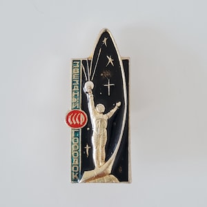 May include: A rectangular metal pin featuring a gold-toned astronaut standing on a base against a black background with stars. The pin includes a red circular emblem with the text CCCP and vertical Cyrillic text reading ZVEZDNY GORODOK.