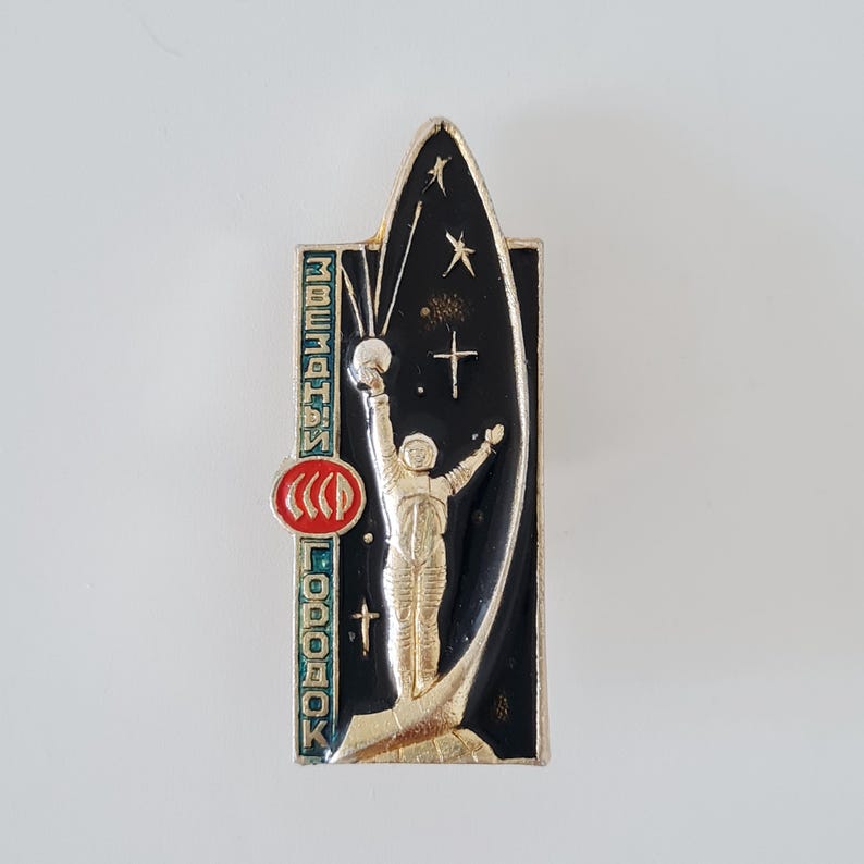May include: A rectangular metal pin featuring a gold-toned astronaut standing on a base against a black background with stars. The pin includes a red circular emblem with the text CCCP and vertical Cyrillic text reading ZVEZDNY GORODOK.