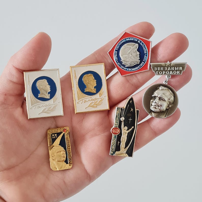 May include: A collection of six metal enamel pins featuring Soviet space exploration themes, including portraits of Yuri Gagarin, rocket imagery, and Cyrillic text. The pins vary in shape, including rectangular, pentagonal, and circular designs.