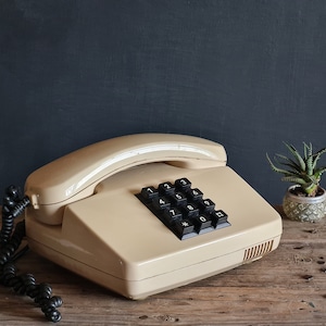 Może przedstawiać: Beżowy telefon w stylu vintage z czarnymi klawiszami numerycznymi i spiralnym kablem. Telefon stoi na rustykalnej drewnianej powierzchni, z małą rośliną doniczkową po prawej stronie i ciemnym tle.