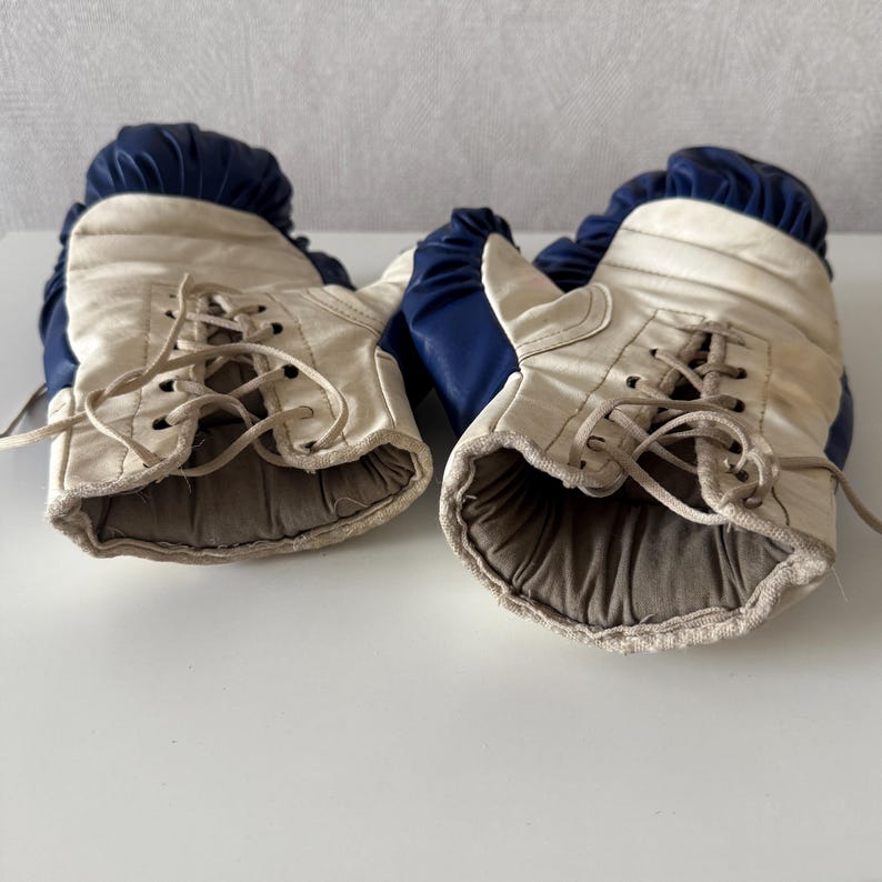 May include: A pair of vintage boxing gloves with white leather and blue trim. The gloves have laces and are worn.