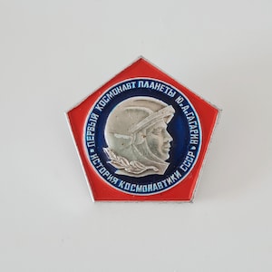 May include: A pentagonal metal pin featuring a red border and a central circular blue field. The center displays a silver-toned relief profile of a cosmonaut in a helmet with Cyrillic text reading "ПЕРВЫЙ КОСМОНАВТ ПЛАНЕТЫ Ю.А.ГАГАРИН ИСТОРИЯ КОСМОНАВТИКИ СССР".