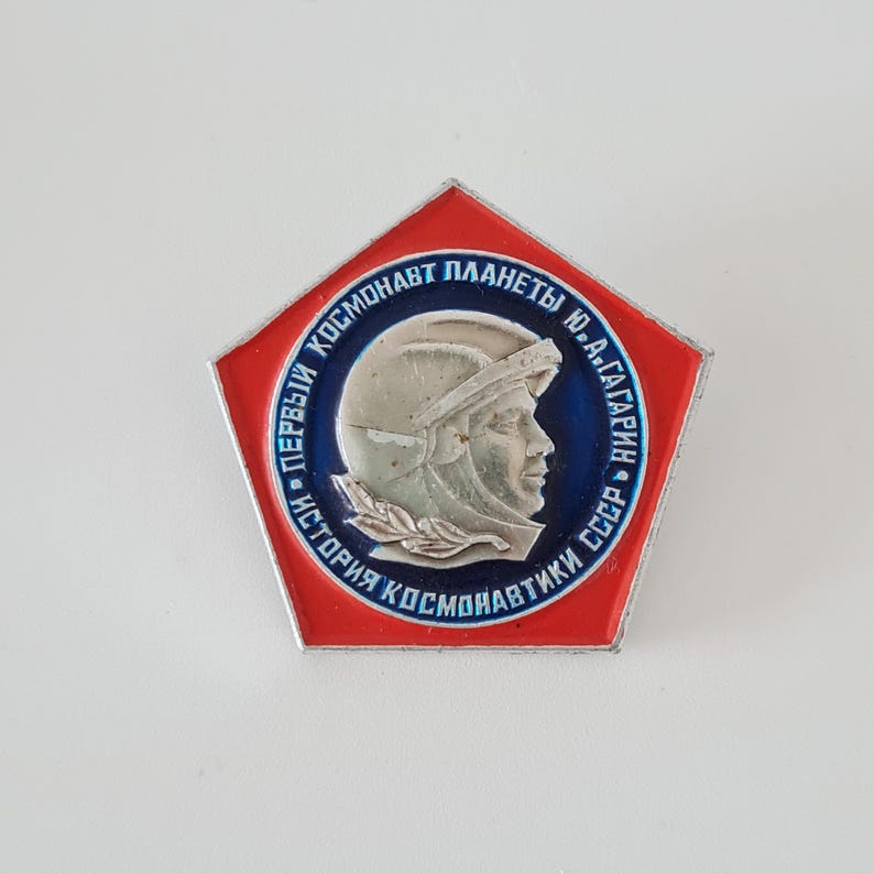 May include: A pentagonal metal pin featuring a red border and a central circular blue field. The center displays a silver-toned relief profile of a cosmonaut in a helmet with Cyrillic text reading "ПЕРВЫЙ КОСМОНАВТ ПЛАНЕТЫ Ю.А.ГАГАРИН ИСТОРИЯ КОСМОНАВТИКИ СССР".