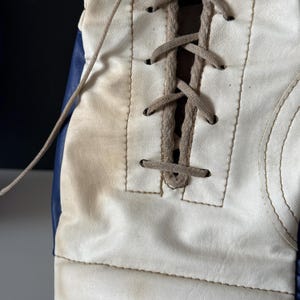 May include: Close-up of a white boxing glove with blue trim and brown laces. The glove is made of leather and has a stitched seam around the edge.
