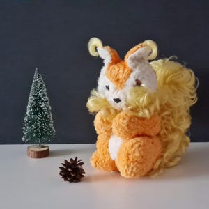 Op de afbeelding: Een pluche oranje en witte eekhoorn met een pluche gele staart. Het speelgoed staat op een wit oppervlak.