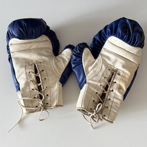 May include: A pair of white boxing gloves with blue trim and lace-up closures. The gloves are worn and show signs of use.