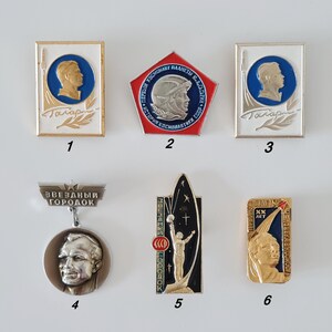 May include: Six Soviet-era space exploration pins featuring Yuri Gagarin and space themes. Pins include rectangular, pentagonal, and circular shapes with metallic finishes, blue, white, and black enamel, and Cyrillic text including Gagarin and CCCP.