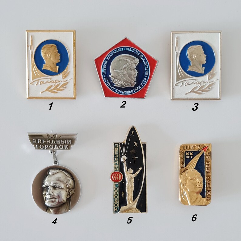 May include: Six Soviet-era space exploration pins featuring Yuri Gagarin and space themes. Pins include rectangular, pentagonal, and circular shapes with metallic finishes, blue, white, and black enamel, and Cyrillic text including Gagarin and CCCP.