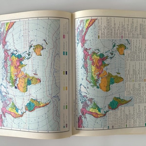 Puede incluir: Libro abierto con dos páginas que muestran mapas mundiales coloridos. Los mapas muestran continentes en tonos verdes, amarillos, naranjas y rosas, con océanos azules y líneas de cuadrícula. Texto visible en la página derecha.