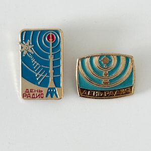 May include: Two vintage gold-toned metal pins. One pin features a tower design with a star and radio wave illustration in blue and white, with the Russian text "Day of Radio." The other pin has a similar design.