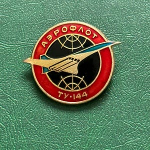 Pode incluir: Um alfinete redondo, dourado, com borda vermelha e centro preto. O alfinete apresenta o texto "АЭРОФЛОТ" em vermelho, um avião estilizado e o texto "ТУ-144" em dourado. O fundo é verde.