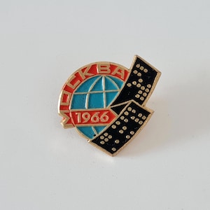 May include: A vintage pin featuring the word "MOSKVA" in red letters, encircling a blue globe with gold accents. The year "1966" is below the globe. A black rectangular shape with white dots is on the right.