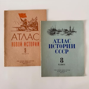Puede incluir: Dos atlas escolares vintage. El atlas de la izquierda tiene una portada marrón con una ilustración de bandera roja y texto en ruso. El atlas de la derecha tiene una portada azul con una ilustración de barco y texto en ruso. Ambos atlas son para clases de historia.
