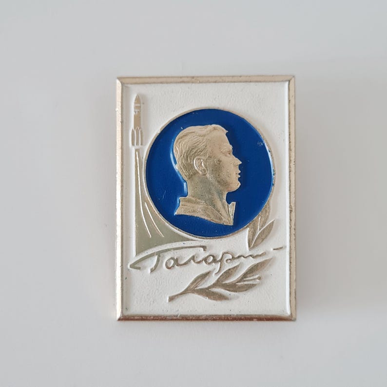 May include: A rectangular metal pin featuring a white background with a silver-toned profile portrait of a person inside a blue circle. A silver rocket and a laurel branch are depicted alongside the Cyrillic text "Гагарин" in silver lettering.