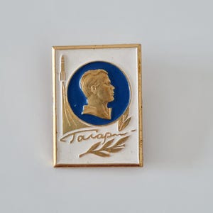 May include: A rectangular metal pin featuring a white background with a gold border. A central blue circle contains a gold profile relief of a person. To the left, a gold rocket ascends, and below, gold Cyrillic text reads Gagarin with a leaf.