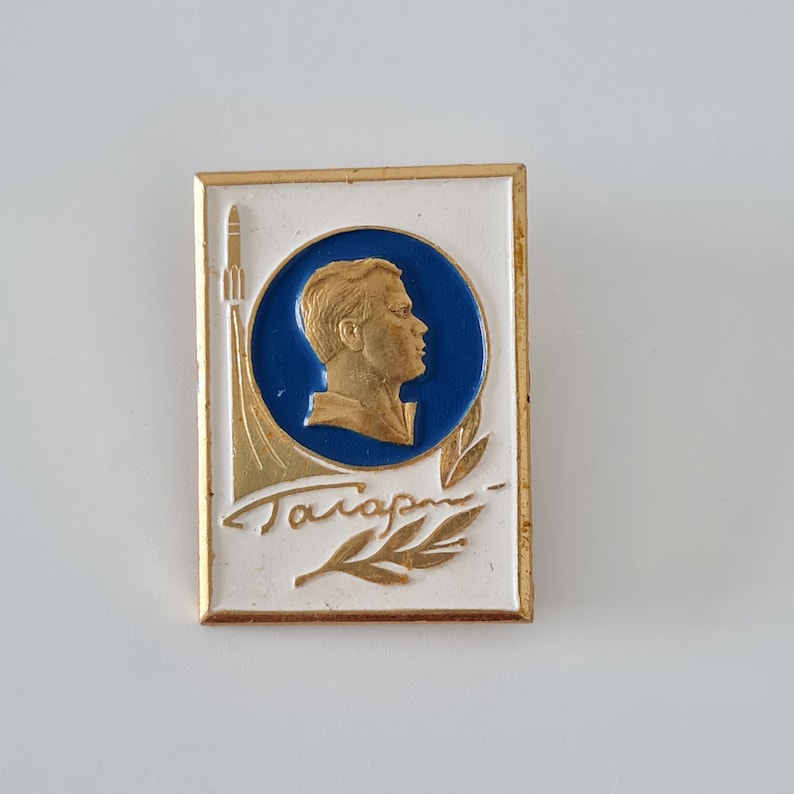 May include: A rectangular metal pin featuring a white background with a gold border. A central blue circle contains a gold profile relief of a person. To the left, a gold rocket ascends, and below, gold Cyrillic text reads Gagarin with a leaf.