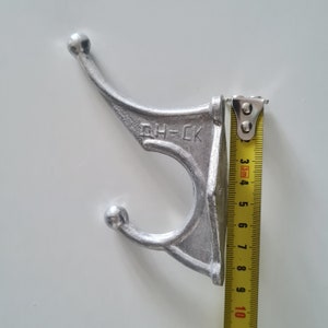 May include: A silver metal hook with two rounded ends and the text "DH-CK" on the side. The hook is being measured with a yellow tape measure.