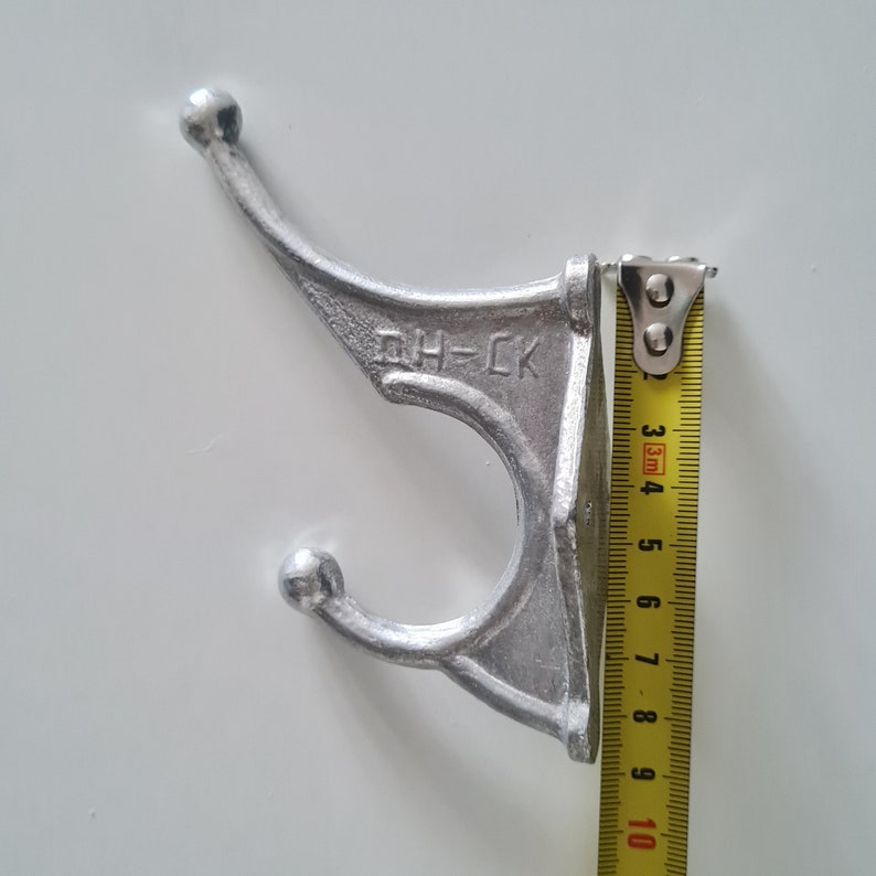 May include: A silver metal hook with two rounded ends and the text "DH-CK" on the side. The hook is being measured with a yellow tape measure.