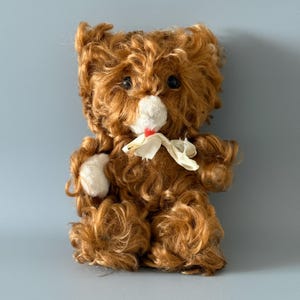 May include: A vintage teddy bear with brown fur, black eyes, and a white muzzle. The bear has a cream-colored bow tied around its neck and white paws. The plush toy is a classic children's item.