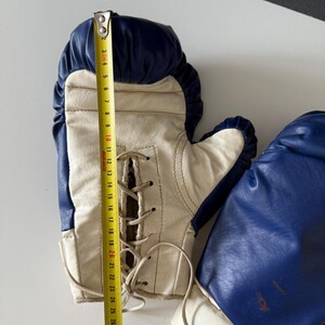 May include: A pair of vintage boxing gloves with white leather and blue accents. The gloves are laced up the back and have a white and blue trim around the wrist.