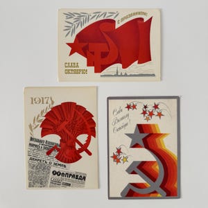 May include: Three vintage postcards with Soviet propaganda imagery. The top card features a red flag with a hammer and sickle, and text in Russian. The middle card has a red wheat sheaf and hammer and sickle over a newspaper. The bottom card has a star and hammer and sickle.