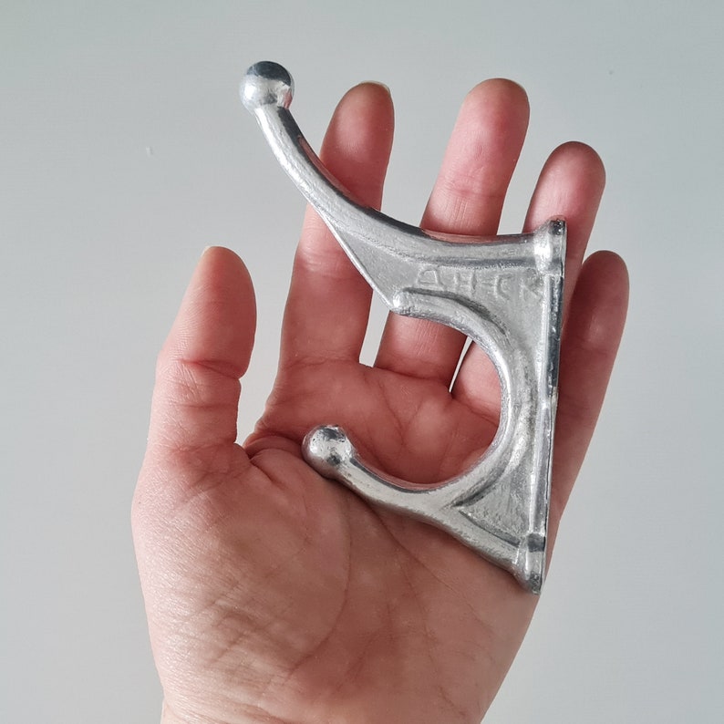 May include: A silver metal double hook with a rounded top and a textured surface. The hook is designed for hanging items on a wall.