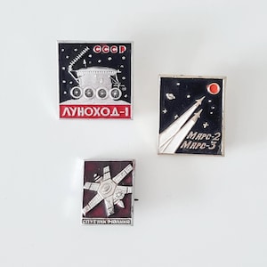 Peut inclure: Trois épingles vintage soviétiques sur le thème de l'exploration spatiale. L'une représente un rover lunaire avec les inscriptions "CCCP" et "LUNOKHOD-1". Une autre montre des fusées et le texte "MARC-2 MARC-3". La troisième représente un satellite avec le texte "SPUTNIK MOLNIYA".