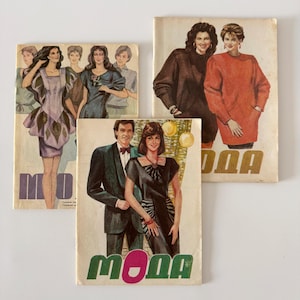 May include: Three vintage fashion magazines with illustrations of people in stylish clothing. The covers feature the word "MODA" in bold lettering. The magazines are on a white background.