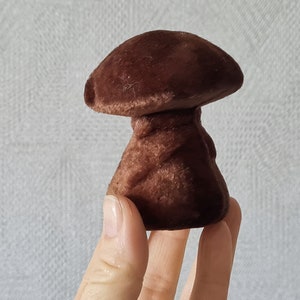 Op de afbeelding: Een bruin, paddenstoelvormig decoratief object met een textuur. De paddenstoel heeft een afgeronde hoed en een steel. Het object wordt in een hand gehouden tegen een witte achtergrond.