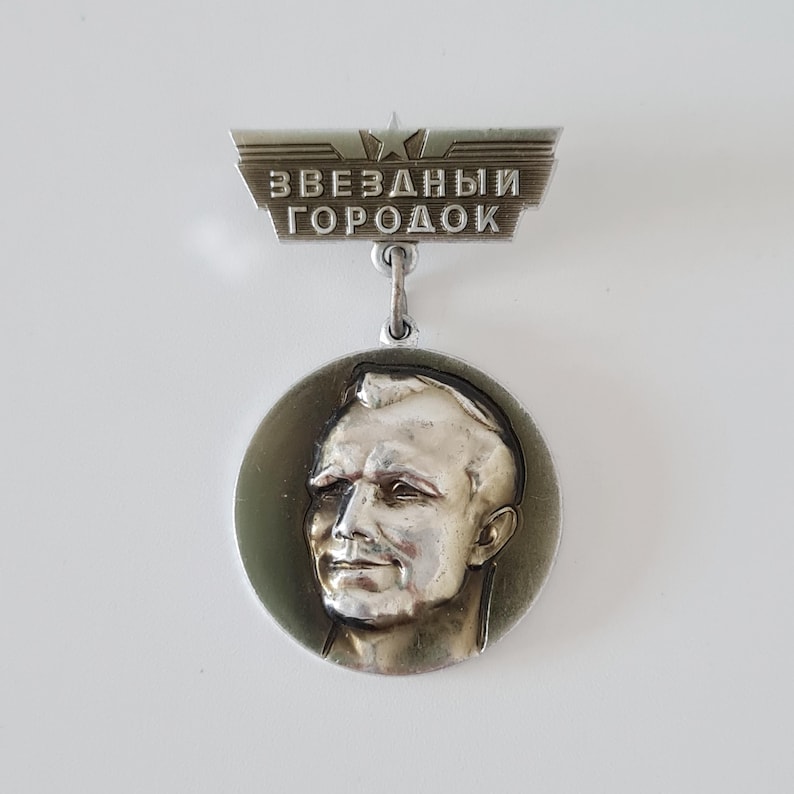 May include: A silver-toned metal commemorative badge featuring a circular pendant with a relief portrait of a person. Above the pendant is a trapezoidal bar with a star and the Cyrillic text ZVEZDNY GORODOK, which translates to Star City.