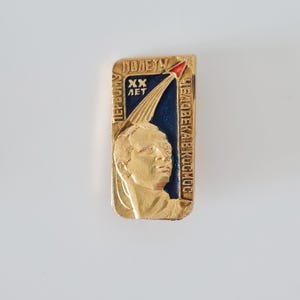 May include: A rectangular gold-toned metal pin featuring a relief portrait of a person, a blue enamel background, and a red-tipped rocket graphic. The pin includes Cyrillic text reading "ПЕРВОМУ ПОЛЕТУ ЧЕЛОВЕКА В КОСМОС" and "XX ЛЕТ" in raised gold lettering.