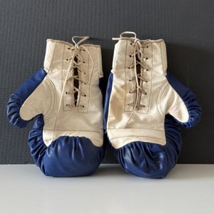 May include: A pair of vintage boxing gloves with white leather and blue padding. The gloves have laces and are tied together.