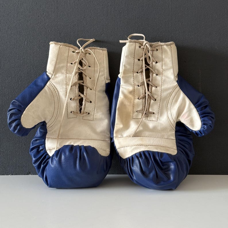 May include: A pair of vintage boxing gloves with white leather and blue padding. The gloves have laces and are tied together.