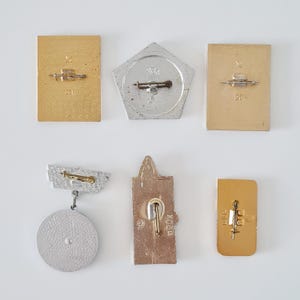 May include: A collection of six metal pins and badges shown from the reverse side. The items include rectangular, pentagonal, and circular shapes in gold and silver tones, each featuring a horizontal pin mechanism for attachment to clothing.
