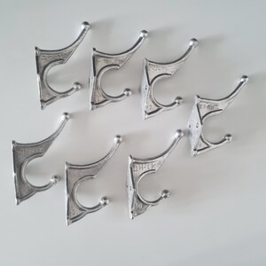 May include: Eight silver metal hooks with a decorative design. Each hook has a rounded top and two prongs. The hooks are arranged in a scattered pattern on a white surface.