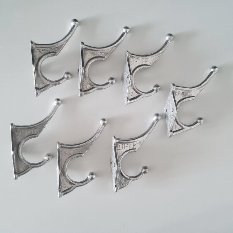 May include: Eight silver metal hooks with a decorative design. Each hook has a rounded top and two prongs. The hooks are arranged in a scattered pattern on a white surface.