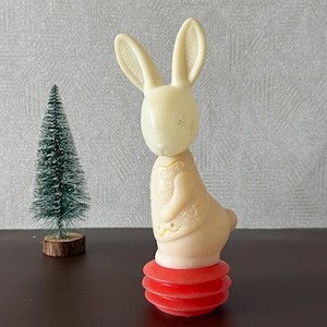 May include: A vintage cream-colored bunny toy with long ears and a red base. The bunny has a detailed design on its body and is standing upright. A small green Christmas tree is in the background.