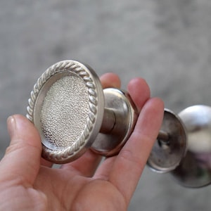 Puede incluir: Candelabros plateados con una parte superior circular texturizada y un borde en forma de cuerda. Los candelabros tienen un tallo y una base redondeada. La imagen muestra tres candelabros sostenidos en una mano.