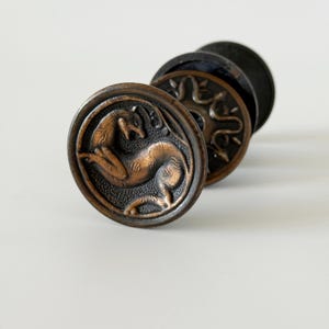 May include: Two circular bronze-toned metal drawer knobs with a dark patina. The front knob features a relief design of a stylized animal in a leaping pose. The second knob behind it displays a circular pattern with radiating leaf-like shapes.