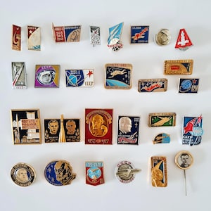 Puede incluir: Colección de insignias y pines vintage de la era soviética sobre exploración espacial. Los pines presentan varios diseños, incluyendo cosmonautas, cohetes y el emblema CCCP. La paleta de colores incluye rojo, dorado, azul y blanco.