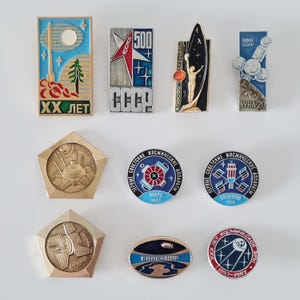 May include: A collection of nine metal space-themed lapel pins featuring various shapes including rectangles, pentagons, circles, and an oval. The pins display Soviet-era imagery, including rockets, satellites, cosmonauts, and Cyrillic text.