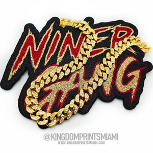 Puede incluir: Un collar dorado con incrustaciones de hielo, con un diseño de cadena cubana gruesa, descansando sobre un parche negro y dorado con las palabras "NINER GANG" en letras rojas y doradas. La imagen incluye el texto "@KINGDOMPRINTSMIAMI" y "WWW.KINGDOMPRINTSMIAMI.COM".