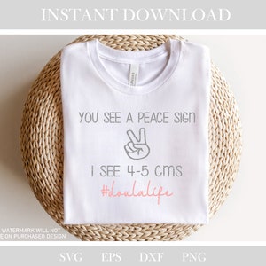 May include: White t-shirt with the text "You see a peace sign, I see 4-5 cms #doulalife" in gray and pink lettering. A peace sign is drawn in gray above the text.