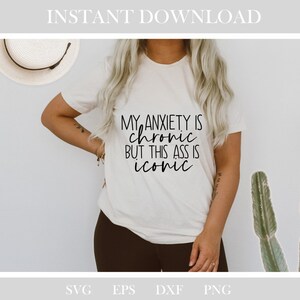 May include: A cream-coloured t-shirt featuring the black text "MY ANXIETY IS chronic BUT THIS ASS IS iconic". The shirt is worn with dark trousers. A straw hat is visible in the background.