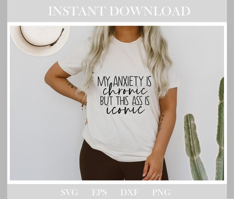 May include: A cream-coloured t-shirt featuring the black text "MY ANXIETY IS chronic BUT THIS ASS IS iconic". The shirt is worn with dark trousers. A straw hat is visible in the background.