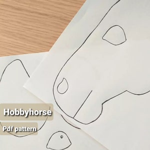 Op de afbeelding: Zwarte omtrek tekeningen van een hobbyhorse hoofd en andere delen op wit papier. De tekst "Hobbyhorse Pdf pattern" staat in een beige rechthoek linksonder. Op een lichtbruin houten oppervlak.